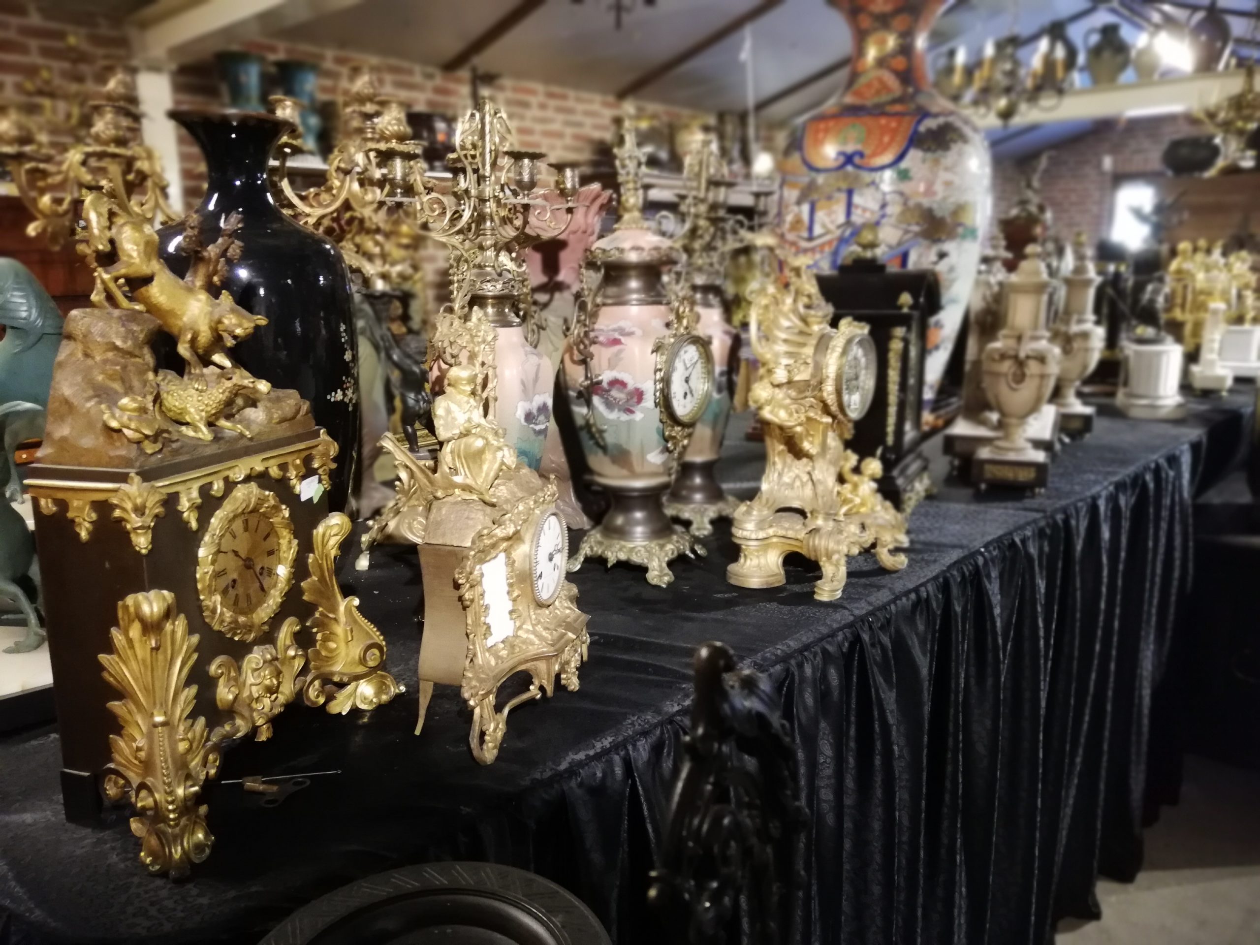 Antieke pendules, kandelaars en vazen in verguld brons op een met zwart fluweel beklede toonbank in de winkel van Baakman Antiques in Gaanderen